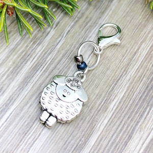 May include: A silver-colored sheep charm with a lobster clasp. The sheep has a smiling face and a fluffy body. The charm is attached to a silver ring and a clasp. The background includes a wooden surface and green pine needles.
