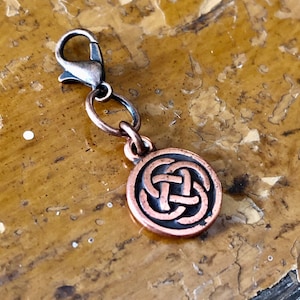 May include: A copper-colored charm with a Celtic knot design. The charm is attached to a lobster clasp. The charm is round and has a raised knot pattern. The clasp and the charm are made of metal.