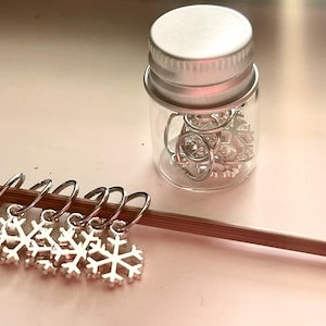 May include: Silver snowflake stitch markers on a wooden knitting needle. A small glass jar with a silver lid contains additional stitch markers. The markers are designed for knitting and crochet projects.