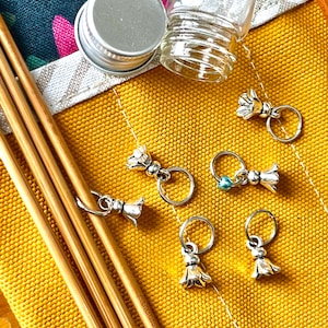 May include: A close-up of a yellow fabric with knitting needles, stitch markers, and a small glass jar with a silver lid. The stitch markers are silver with a flower design. The knitting needles are bamboo.