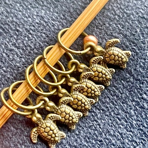 May include: A collection of antique gold-colored turtle stitch markers attached to a bamboo knitting needle. Each marker features a detailed turtle charm. A small, peach-colored bead is also present.