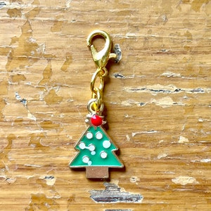 May include: A gold-colored Christmas tree charm with a lobster clasp. The green tree has white dots and a red ornament at the top. The charm is attached to a gold chain.