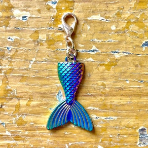 Puede incluir: Un dije de cola de sirena con cierre de mosquetón. El dije presenta un acabado iridiscente vibrante con tonos de azul, verde y morado. La cola está detallada con patrones de escamas y aletas.