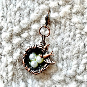 May include: A bronze-colored charm featuring a bird's nest with three pearl-like beads and a small bird. The charm is attached to a clasp, suitable for jewelry or craft projects. The nest has a dark green interior.