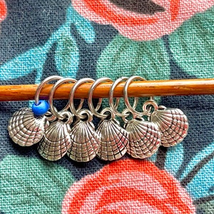 May include: Silver-toned seashell stitch markers with a blue bead, attached to a wooden knitting needle. The markers are designed to help track rows or stitches in knitting projects. The background is a floral fabric.
