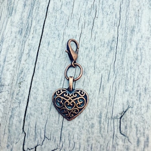 Puede incluir: Un dije en forma de corazón de color cobre con un intrincado diseño calado, suspendido de un cierre de mosquetón y un pequeño anillo. El dije es probablemente para un collar o pulsera, y está sobre un fondo de madera envejecida.