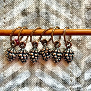 Puede incluir: Seis marcadores de punto de color bronce con forma de piña están unidos a una aguja de tejer de madera. Cada marcador tiene un pequeño lazo y un diseño detallado de piña. Se ve una sola cuenta roja. El fondo es una tela clara y texturizada.