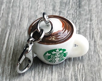 May include: A miniature coffee cup charm with a silver clasp. The cup is white with a green logo and filled with a brown liquid and a swirl of white. The charm is designed to resemble a popular coffee shop's beverage.