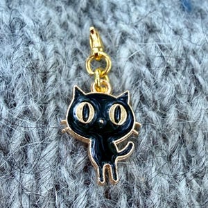 Puede incluir: Un dije de metal en tono dorado con forma de gato negro, grandes ojos amarillos separados y un acabado de esmalte brillante. El dije cuenta con un pequeño cierre de mosquetón dorado en la parte superior, unido por una argolla circular.