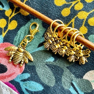 Puede incluir: Marcadores de punto y un dije dorados con temática de abeja. El dije es un colgante de abeja detallado con un cierre de mosquetón. Los marcadores de punto son pequeños anillos con adornos de abeja. Los artículos descansan sobre una aguja de tejer de madera, sobre un fondo de tela floral.