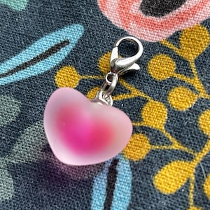 May include: A heart-shaped charm with a silver clasp. The charm is a gradient of pink, with a lighter pink on the outside and a deeper pink in the center. The charm is likely made of glass or a similar material.