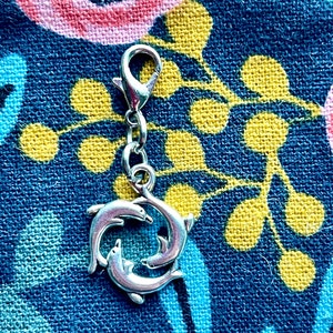 May include: A silver charm with a clasp, featuring three dolphins in a circular arrangement. The charm is designed for jewellery. The background is a blue fabric with floral patterns.