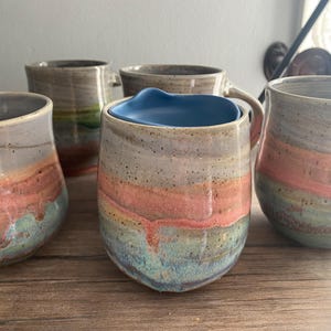 May include: A collection of handmade ceramic mugs with a unique, layered design. The mugs feature bands of color in shades of gray, coral, and teal. One mug has a blue interior. The mugs are on a wooden surface.