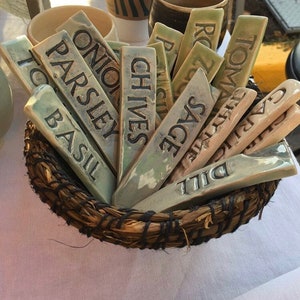 May include: A wicker basket filled with ceramic plant markers. The markers are labeled with the names of herbs, including Basil, Parsley, Onion, Chives, Sage, Dill, Garlic, Oregano, Rosemary, and Thyme.