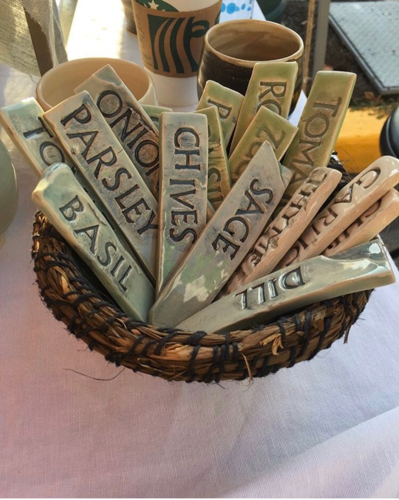 May include: A wicker basket filled with ceramic plant markers. The markers are labeled with the names of herbs, including Basil, Parsley, Onion, Chives, Sage, Dill, Garlic, Oregano, Rosemary, and Thyme.