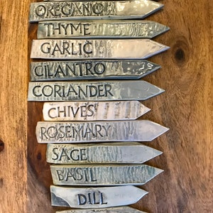 May include: A set of 12 ceramic garden markers with black lettering. The markers are in shades of gray and white and feature the names of common herbs: Parsley, Oregano, Thyme, Garlic, Cilantro, Coriander, Chives, Rosemary, Sage, Basil, Dill, and Mint.