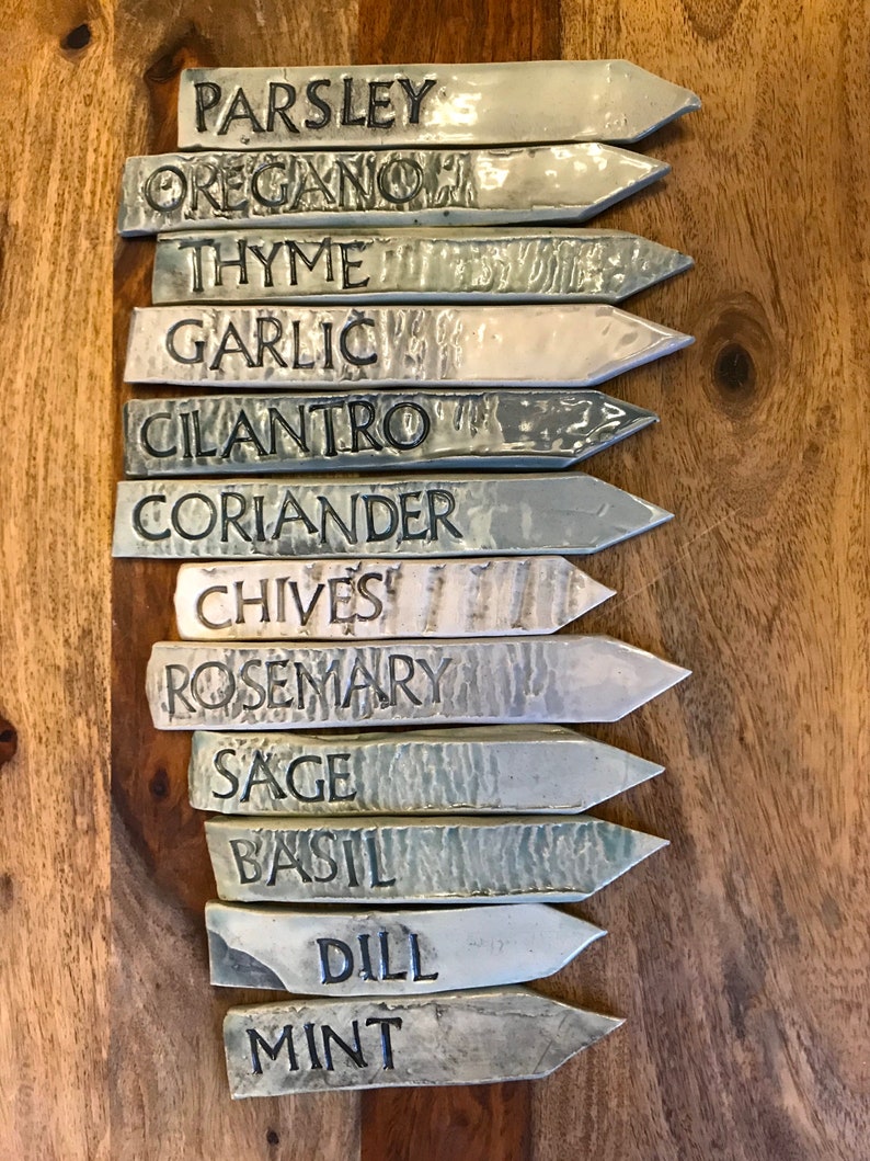 May include: A set of 12 ceramic garden markers with black lettering. The markers are in shades of gray and white and feature the names of common herbs: Parsley, Oregano, Thyme, Garlic, Cilantro, Coriander, Chives, Rosemary, Sage, Basil, Dill, and Mint.