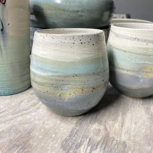 May include: A collection of handmade ceramic drinkware. The pieces include a tall, cylindrical vase and several rounded, stemless glasses and bowls. The pottery features a layered design with shades of white, blue, and yellow, with speckled detailing.