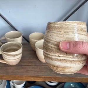 May include: A hand holds a beige and brown speckled ceramic tumbler. The tumbler has a rounded base and a cylindrical top. Other ceramic mugs and tumblers are on a wooden shelf in the background.