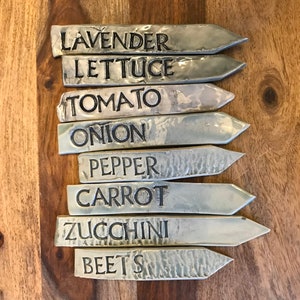 May include: Set of eight silver-toned metal garden markers with black lettering. The markers are labeled: Lavender, Lettuce, Tomato, Onion, Pepper, Carrot, Zucchini, and Beets.