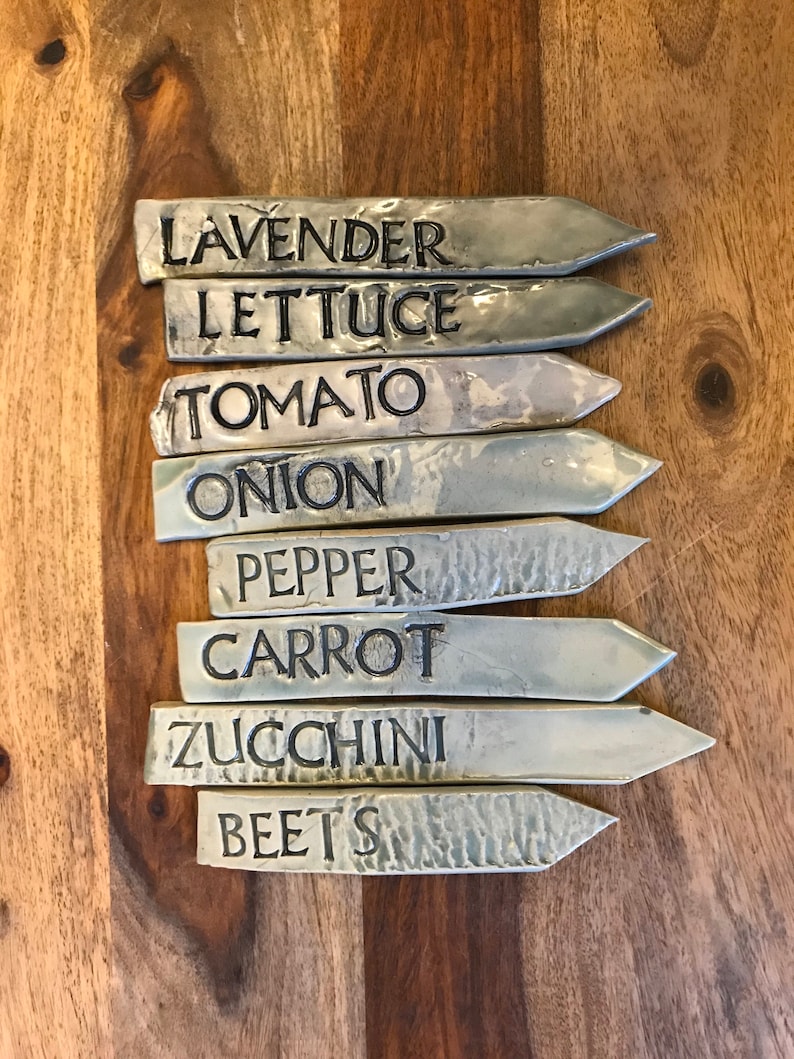 May include: Set of eight silver-toned metal garden markers with black lettering. The markers are labeled: Lavender, Lettuce, Tomato, Onion, Pepper, Carrot, Zucchini, and Beets.