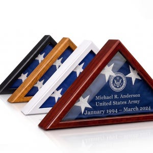 May include: Four triangular flag display cases in black, oak, white, and cherry wood finishes. Each case holds a folded American flag. The cherry case features engraved text: Michael R. Anderson, United States Army, January 1994 – March 2024.