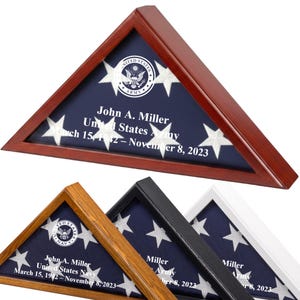 May include: A wooden triangular flag display case with a blue background and white stars. The case contains a folded American flag and text that reads "John A. Miller, United States Army, March 15, 1942 - November 8, 2023". Additional cases in different colors are visible.