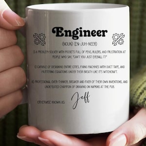 May include: White ceramic mug with the word "Engineer" in a bold, stylized font. The mug features a humorous definition of an engineer, including phrases like "problem-solver" and "fixer of their own inventions." The name "Jeff" is written below.