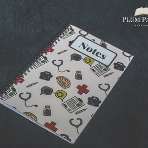 May include: A white spiral notebook with a colorful pattern of medical symbols and the word "Notes" on the front cover. The notebook is on a dark gray surface.