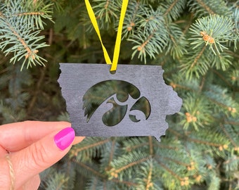 May include: Black wooden ornament shaped like the state of Iowa with a cutout of the University of Iowa Hawkeye logo. The ornament is hanging from a yellow ribbon on a green pine tree.