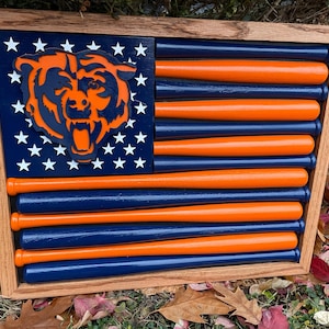 May include: Wooden wall art featuring a Chicago Bears-themed American flag design. The flag incorporates a bear head, white stars, and alternating blue and orange baseball bats, all set within a wooden frame.