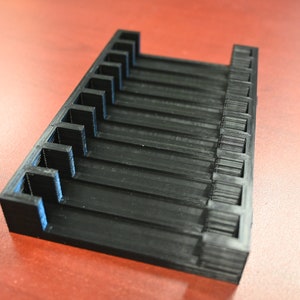 May include: A black plastic organizer with 12 slots for storing small items. The organizer is made of 3D printed plastic and has a smooth, matte finish.