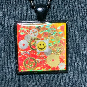 May include: A square pendant necklace with a red, orange, and yellow background. The pendant is filled with gold and silver gears and a yellow smiley face sticker.