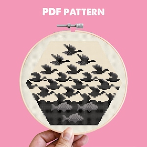 Può includere: Un ricamo a punto croce in un telaio di legno con un disegno in bianco e nero ispirato a Escher di uccelli e pesci. Il testo "PDF PATTERN" è in alto. Il ricamo è tenuto da una mano su uno sfondo rosa.