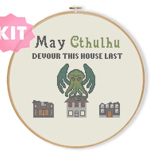 Peut inclure: Un kit de point de croix avec un tambour à broder en bois contenant un tissu blanc avec le texte May Cthulhu DEVOUR THIS HOUSE LAST en noir et vert, une figure de Cthulhu verte et trois petites maisons grises brodées en bas.