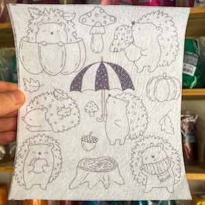 Peut inclure: Un tissu blanc avec une illustration en noir et blanc de hérissons, de champignons, d'un parapluie, d'une citrouille et d'une souche d'arbre. Les hérissons sont dans diverses poses, certains tenant des parapluies, d'autres portant des citrouilles et d'autres dormant sous des champignons.