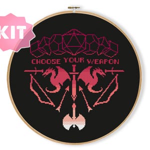 May include: Cross-stitch kit with a black background and a red and pink design. The design includes geometric shapes, dragons, swords, and the text "CHOOSE YOUR WEAPON". The kit is in a wooden hoop with a pink "KIT" label.