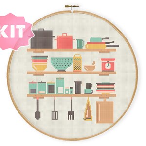 Könnte beinhalten: Kreuzstich-Set mit einer Küchenszene mit Regalen, die Küchenutensilien halten. Das Design umfasst Töpfe, Pfannen, Schüsseln, eine Reibe und einen Mörser mit Stößel. Das Wort "KIT" befindet sich in einem rosa Stern.