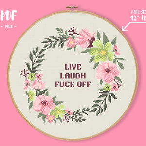 May include: A cross-stitch hoop with a floral wreath design. The wreath features pink and green flowers and dark green leaves. The text "LIVE LAUGH FUCK OFF" is stitched in the centre. The hoop is 30.5 cm in diameter.