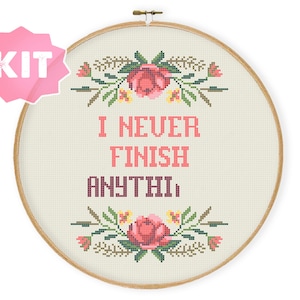 May include: Cross-stitch kit in a wooden hoop with the text "I NEVER FINISH ANYTHING" in pink and burgundy. The design includes floral accents in pink, green, and yellow. A pink starburst-shaped label reads "KIT".