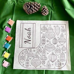 Könnte beinhalten: Ein weißer Weihnachtsstrumpf mit einem schwarzen Umrissdesign von Ornamenten und dem Namen "Noah". Das Bild zeigt Garnrollen und Tannenzapfen auf einer grünen Oberfläche.