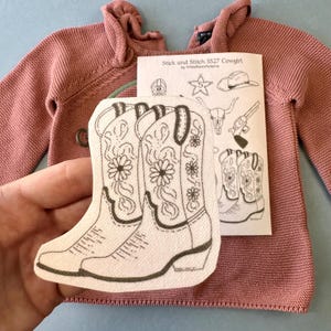 May include: A pink knitted hooded sweater with a matching iron-on patch featuring a pair of cowboy boots with floral designs. A paper with the text "Stick and Stitch SS27 Cowgirl" is also visible.
