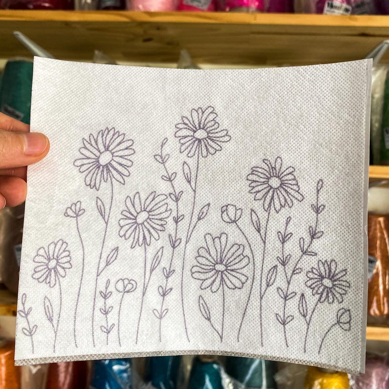 May include: A white fabric with a design of purple flowers and stems. The flowers are simple and have a daisy-like shape.
