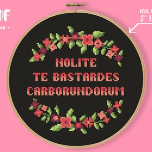 May include: Black and white cross-stitch pattern with a floral wreath and the text "Holite Te Bastardes Carborundum". The pattern is displayed in an 8-inch embroidery hoop.