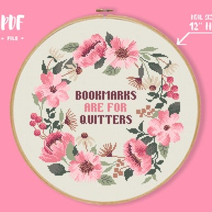May include: A cross-stitch design in a 12-inch hoop features a floral wreath of pink flowers and green leaves. The text "BOOKMARKS ARE FOR QUITTERS" is centered within the wreath. The background is a light beige fabric.