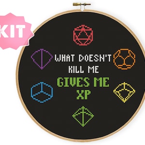 Puede incluir: Un aro de bordado negro con un diseño de punto de cruz que presenta dados coloridos y el texto "What doesn't kill me gives me XP".
