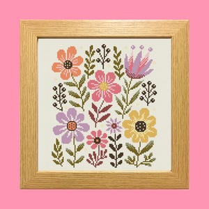 Pode incluir: Um design floral em ponto cruz, emoldurado, com uma variedade de flores coloridas em tons de laranja, rosa, roxo e amarelo. As flores são dispostas com folhas e caules verdes sobre um fundo creme. A moldura é de madeira clara.