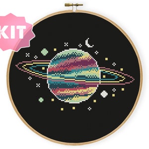 Puede incluir: Un kit de punto de cruz con un planeta colorido con anillos, una luna creciente y estrellas sobre un fondo negro. El kit incluye todos los materiales necesarios para completar el proyecto.