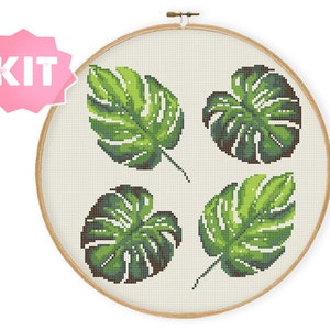 May include: A cross-stitch embroidery kit featuring four green monstera leaves on a white background. The kit is displayed in a wooden embroidery hoop.
