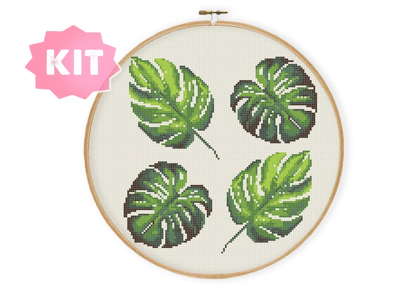 May include: A cross-stitch embroidery kit featuring four green monstera leaves on a white background. The kit is displayed in a wooden embroidery hoop.